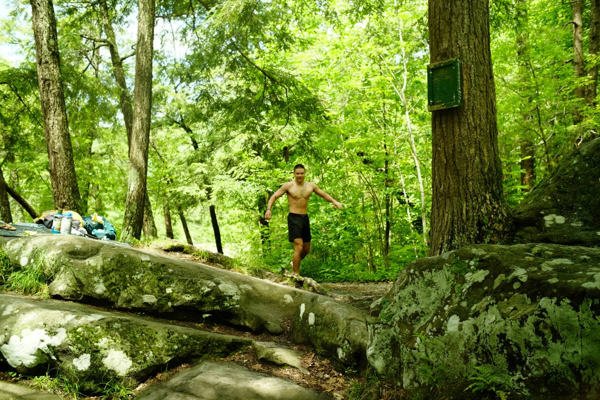 A person wearing hiking boots stands atop a hill overlooking a forest at sunrise.