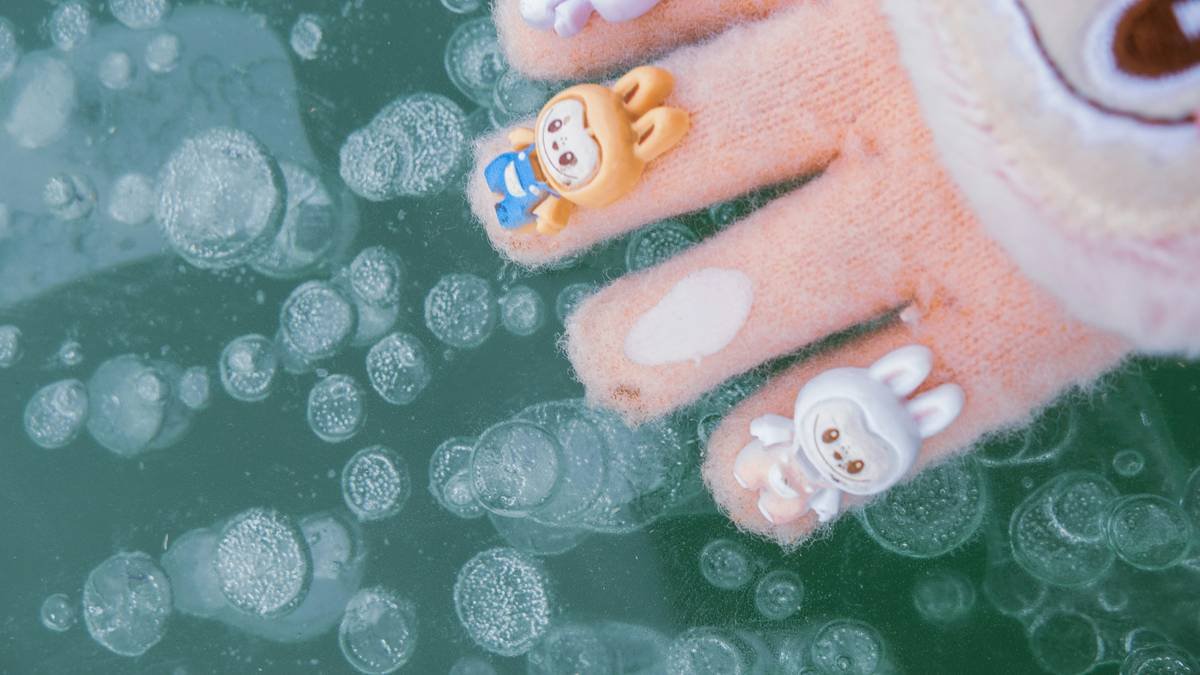 Close-up of a fuzzy peach-colored glove with cute cartoon character finger puppets, resting above a turquoise icy surface filled with frozen bubbles—whimsical winter detail with playful charm.